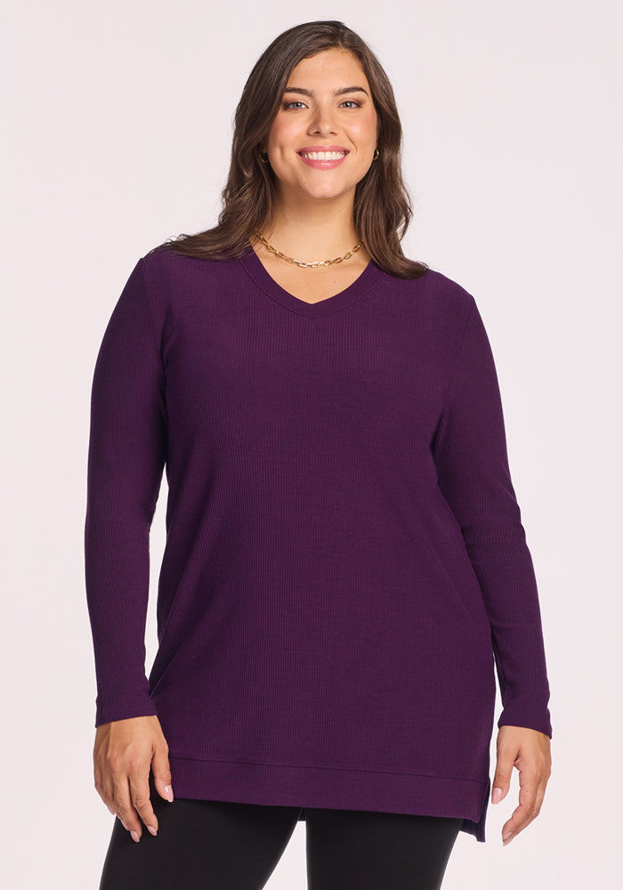 A woman with long brown hair smiles in the Woolx Evie Ribbed Tunic in Majestic Purple, a cozy Merino wool sweater, paired with a gold chain necklace and black pants, standing against a plain light background. 