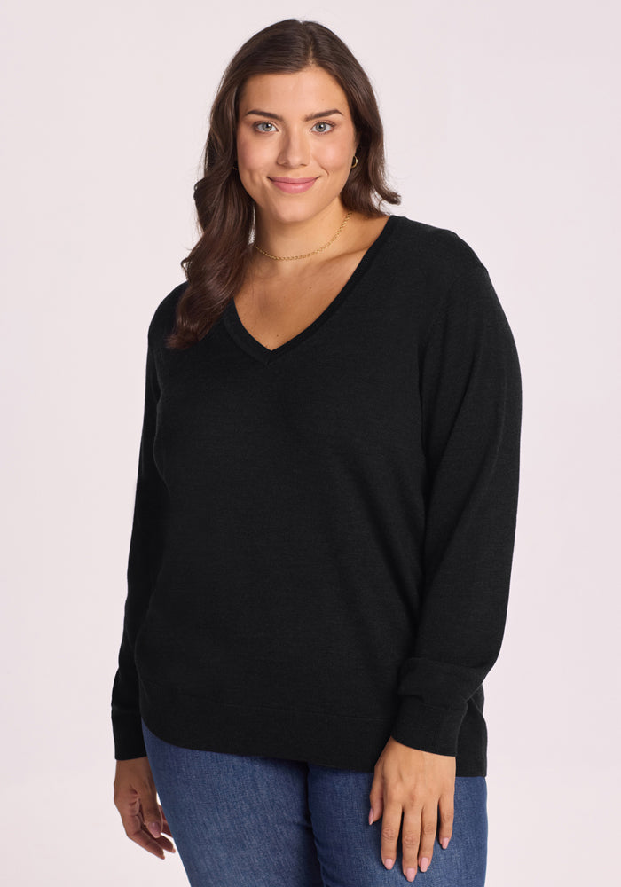 A woman with long brown hair smiles softly at the camera, wearing the travel-friendly Woolx Francesca Sweater in black and blue jeans, standing against a plain light background. 
