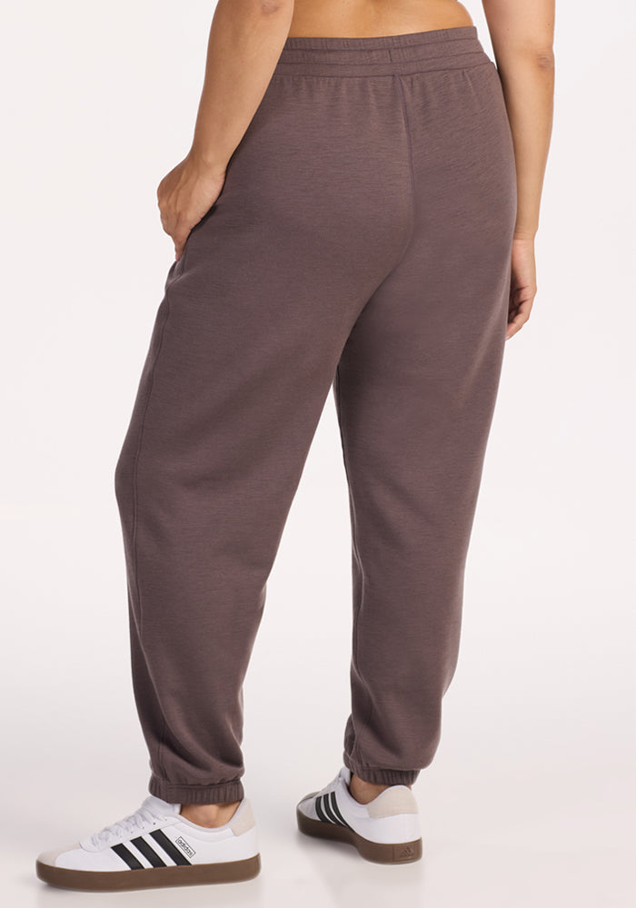 A person, seen from behind against a plain light background, wears relaxed-fit Woolx Parker Sweatpants in Plum Truffle—crafted from Merino wool for moisture-wicking and temperature regulation—paired with white Adidas sneakers with black stripes.