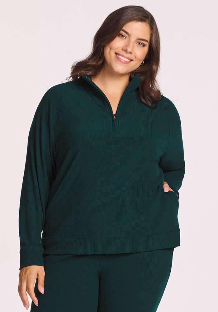 A woman with long brown hair smiles while wearing the Woolx Quinn Ribbed 1/4 Zip in Deep Timber and matching pants, standing against a plain light background. 