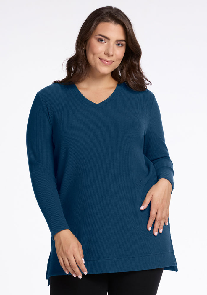A woman with long brown hair wears the Woolx Evie Ribbed Tunic in Majolica Blue and black pants, smiling against a plain white background. 