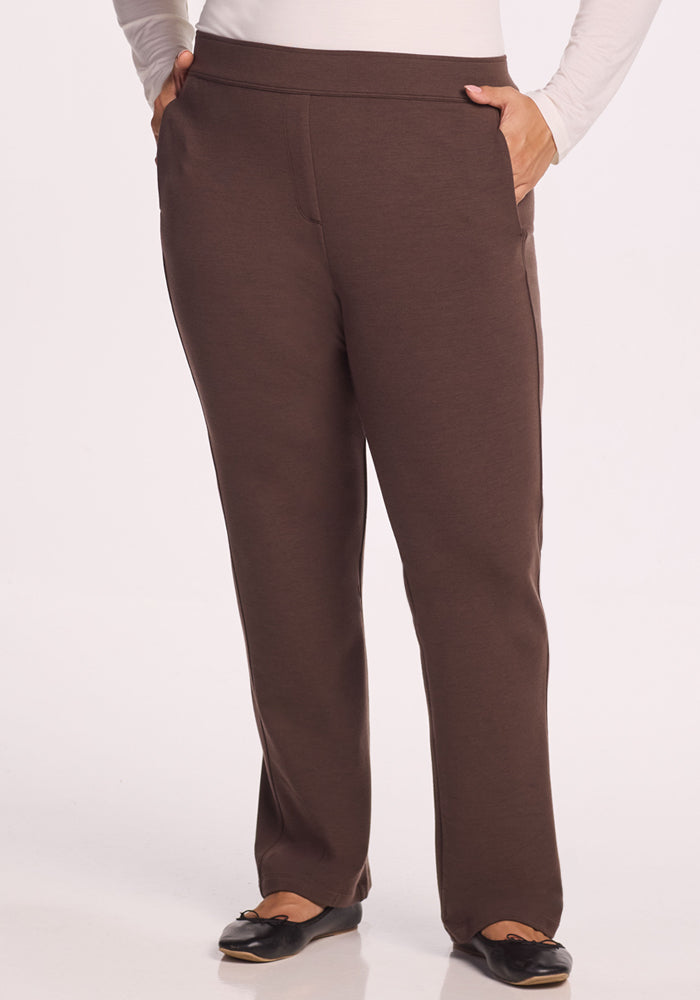 A person wearing Woolx Emerson Straight Leg Pants in Bracken and a white long-sleeve top, with hands in their pockets and black flats, stands against a plain background. 