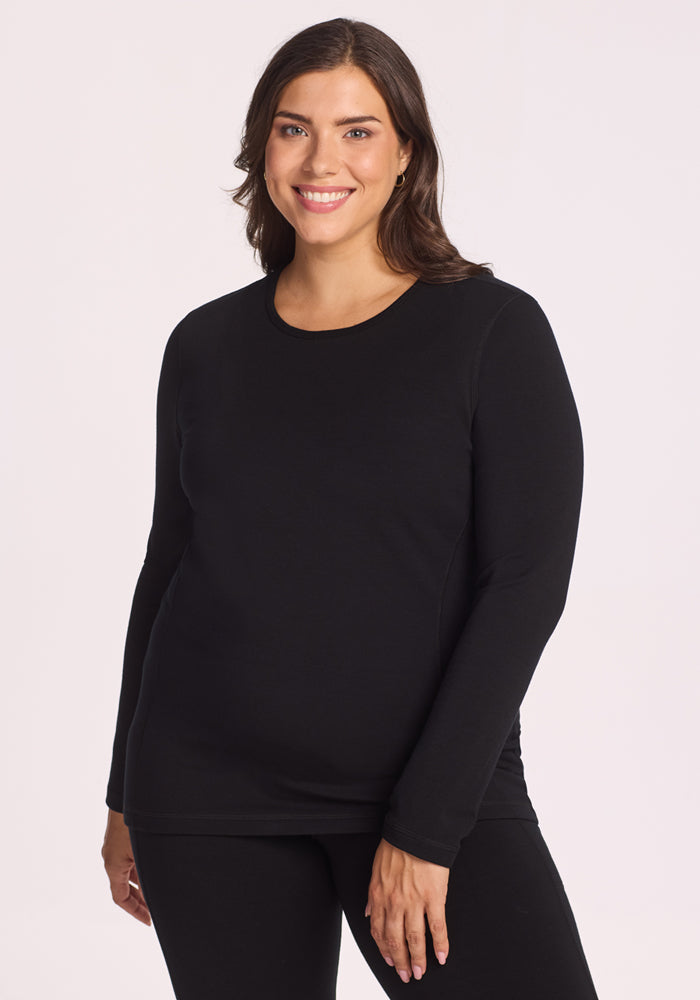 A smiling woman with medium-length brown hair models the Woolx Elsa Long Sleeve in black, paired with matching black pants, standing against a plain light background. 
