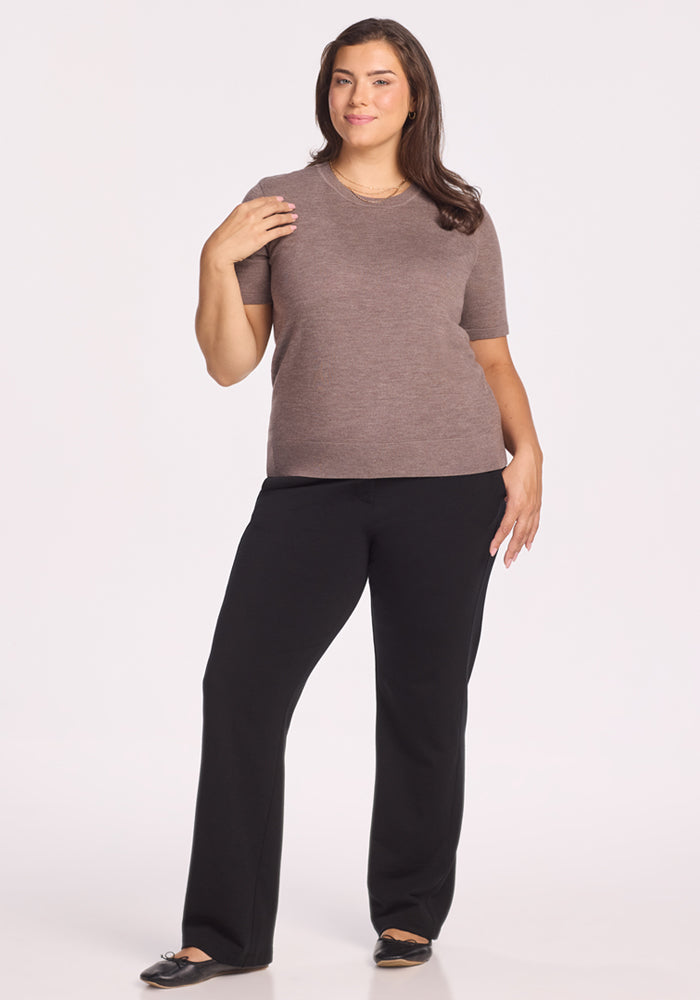 A woman with long brown hair smiles gently against a plain background, wearing the Woolx Anna Crew Sweater in Simply Taupe, black pants, and black flats. She poses confidently with one hand touching her neck.
