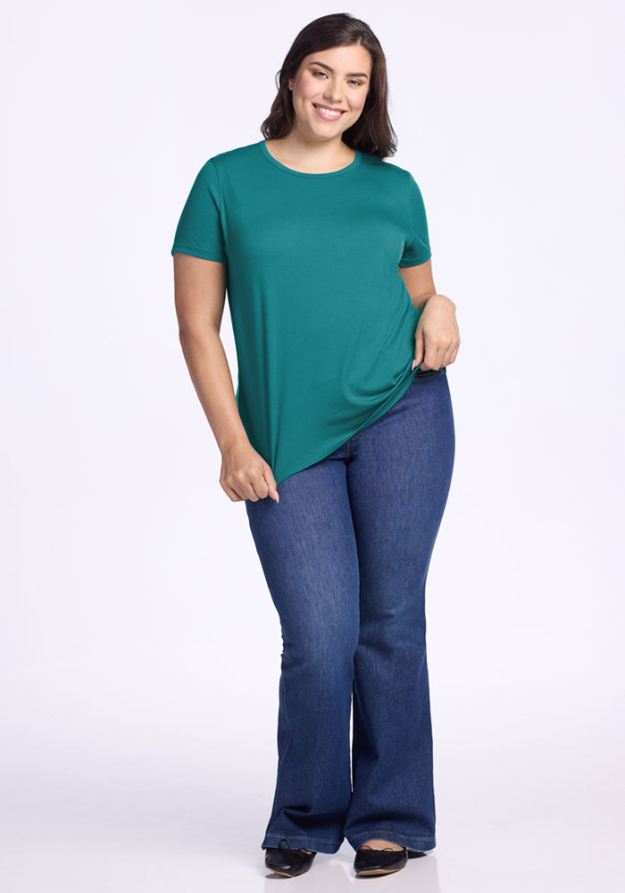 A smiling woman poses confidently against a white background, wearing Woolx's Liza Crew Neck Tee in Surfside and blue flared jeans with black shoes, holding one side of her fitted green top.