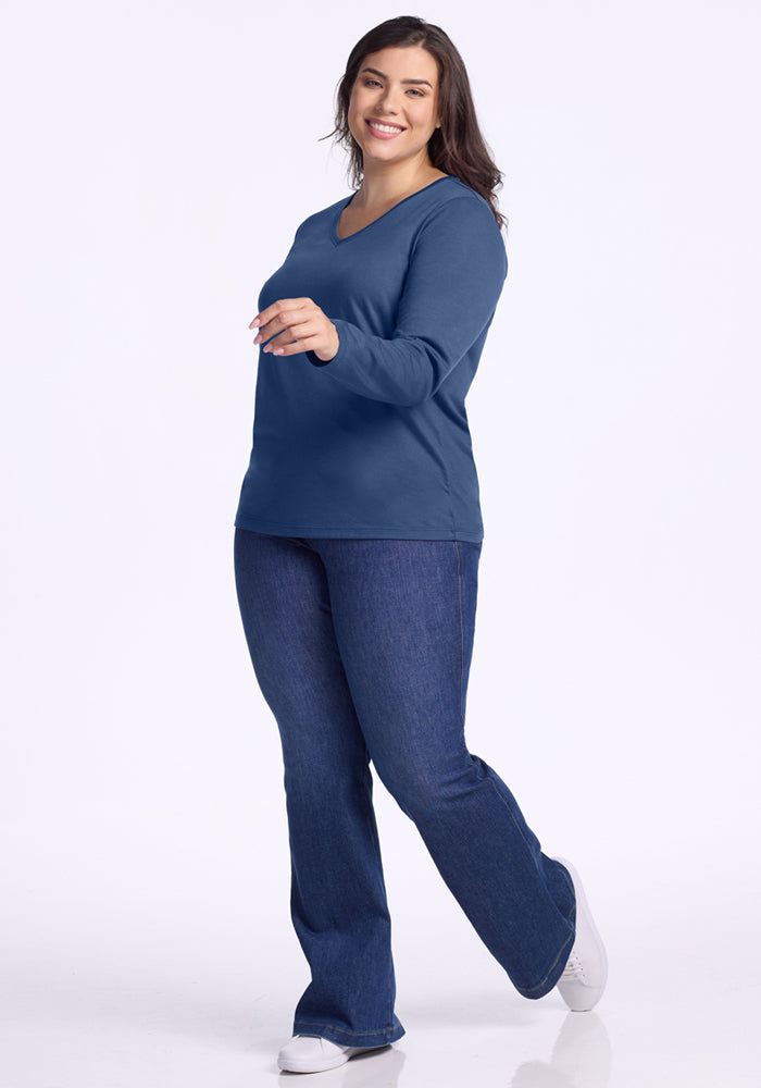 A smiling woman with long dark hair wears the Woolx Millie Long Sleeve - Starry Night, blue jeans, and white sneakers. Standing on one foot and turned to the side, she stays comfortable in the temperature regulating top as she poses gracefully.