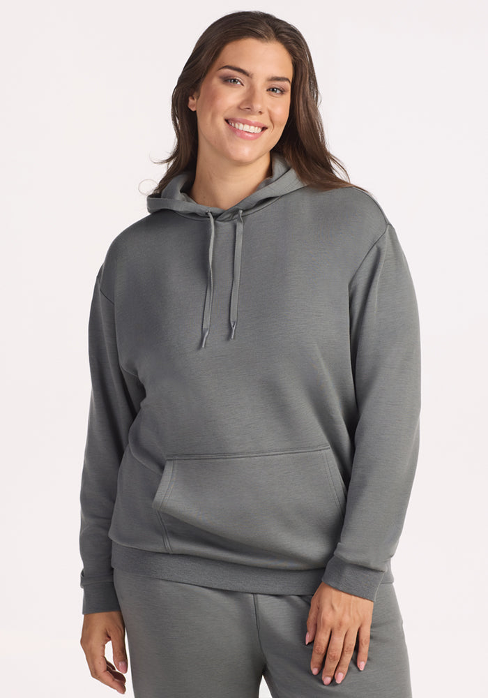 A woman with long brown hair smiles while wearing the Woolx Avery Hoodie in Sedona Sage—an antimicrobial, loose-fitting top—paired with matching sweatpants, standing against a plain white background. 