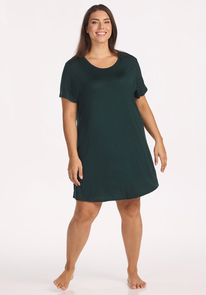 A woman with long brown hair stands barefoot, smiling, and wearing the Woolx Desi Nightgown in Deep Timber—a loose-fitting, short-sleeved Merino wool nightgown—perfect for odor-resistant, cooling sleepwear—against a white background. 