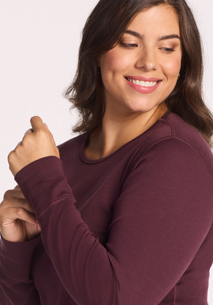 A woman with medium-length brown hair smiles and looks to the side, wearing the Woolx Hannah Long Sleeve Top in Chocolate Fig, a moisture-wicking and temperature-regulating merino wool piece. She pairs it with a natural makeup look.