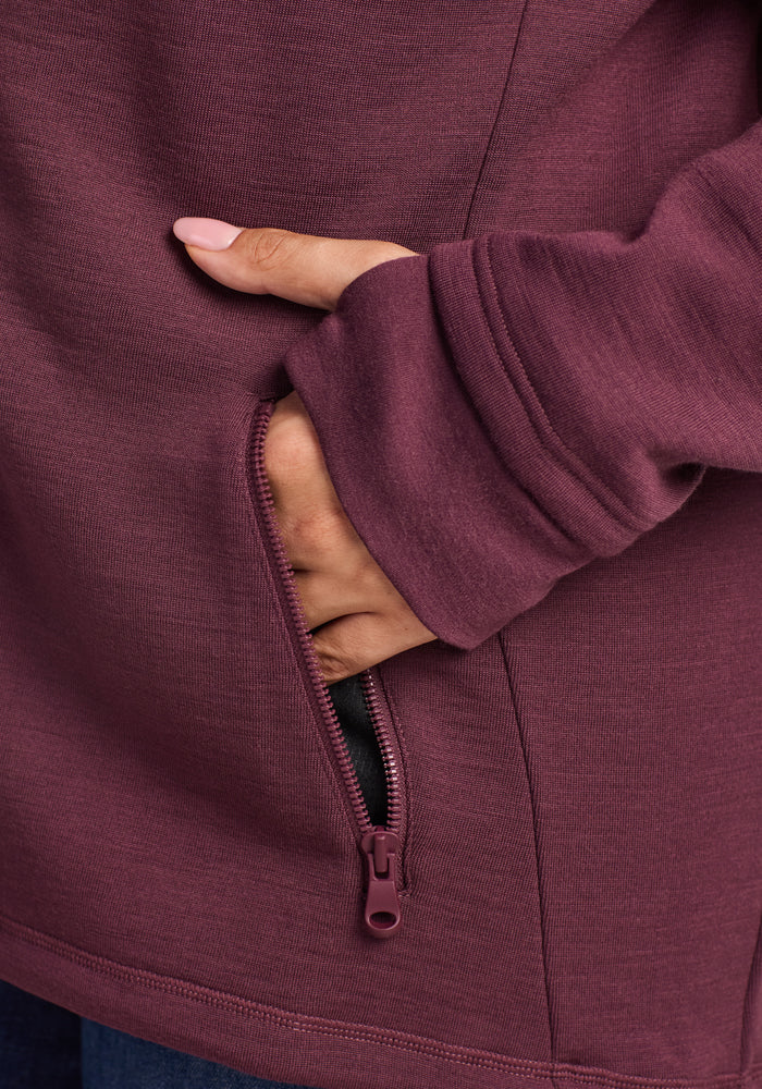 A person’s hand, with light pink nails, partially in a zippered pocket on the maroon Woolx Cubby Hooded Sweatshirt – Grape Harvest.