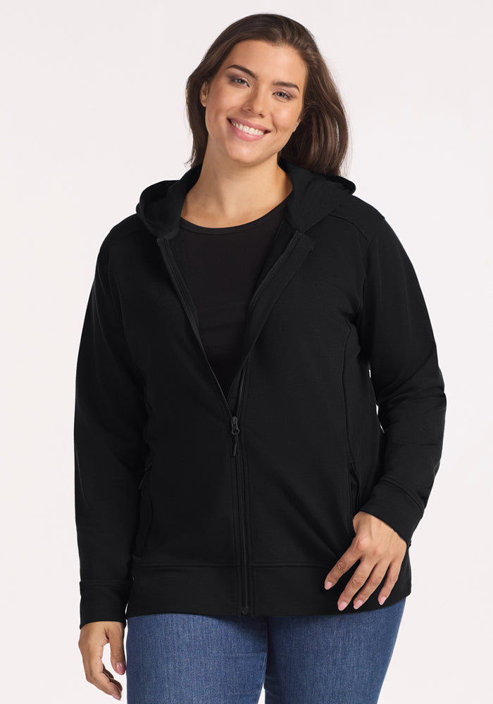 A woman with long brown hair smiles wearing the Woolx Darby Zip Up Hoodie in black, paired with a black shirt and blue jeans, standing against a plain light background. 