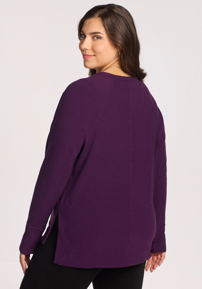 A woman with long brown hair wears the Woolx Ainsley Sweater in Majestic Purple and black pants, standing against a plain light background while smiling slightly over her shoulder.