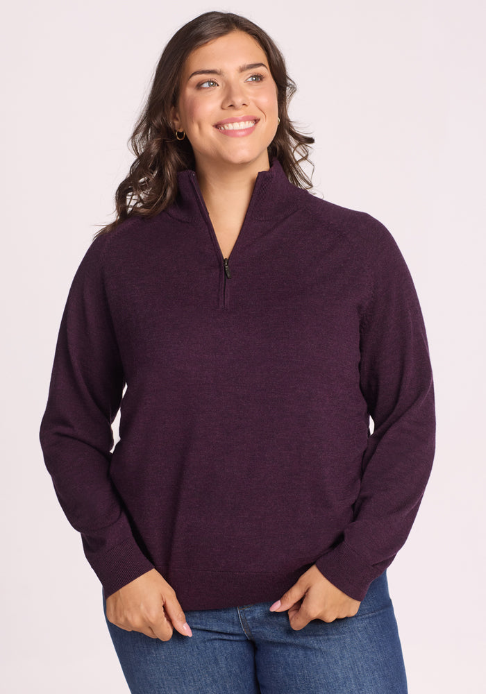 A woman with long brown hair, wearing the Woolx Collins Half Zip Sweater in Deep Plum and blue jeans, stands smiling with her hands relaxed in front of her against a plain light background. 