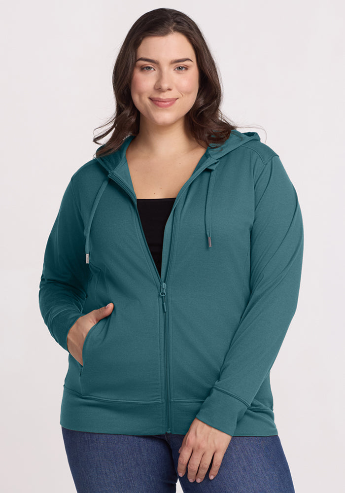 A woman with long brown hair smiles with one hand in her pocket, wearing the Woolx Ryann Hoodie in Tidepool over a black top and blue jeans, standing against a plain light background. 