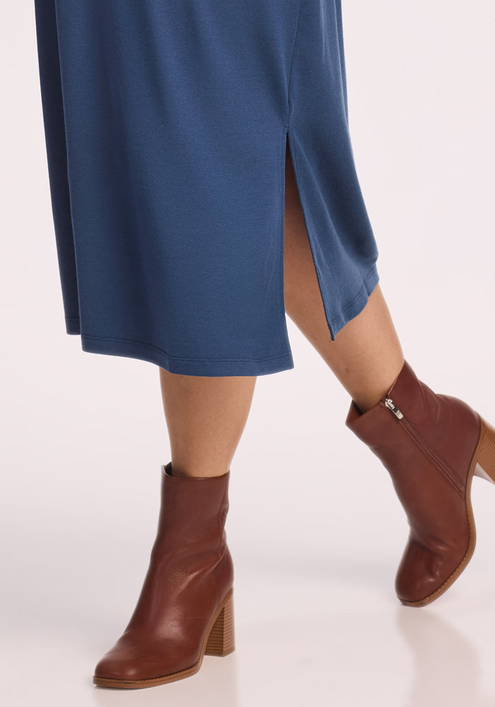 Someone wearing the Woolx Cassie Dress in Starry Night, featuring moisture-wicking fabric and a side slit, paired with brown heeled ankle boots, walking on a light background.