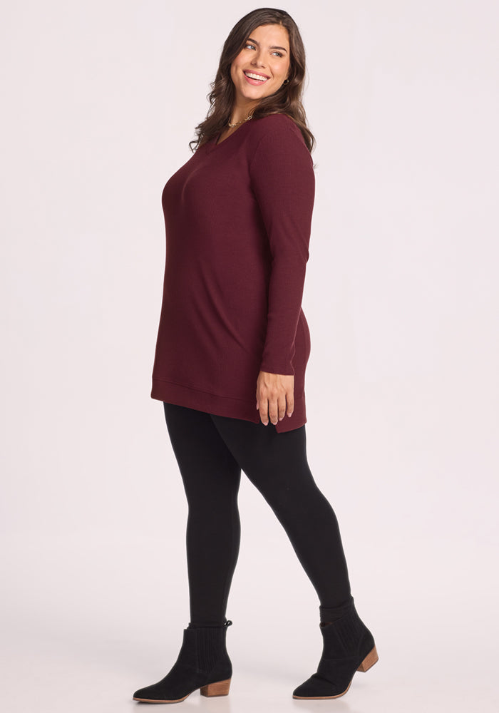 A woman with long dark hair poses sideways, smiling in a cozy Woolx Evie Ribbed Tunic in Toasted Garnet, paired with black leggings and low-heeled black ankle boots. The background is plain light gray.