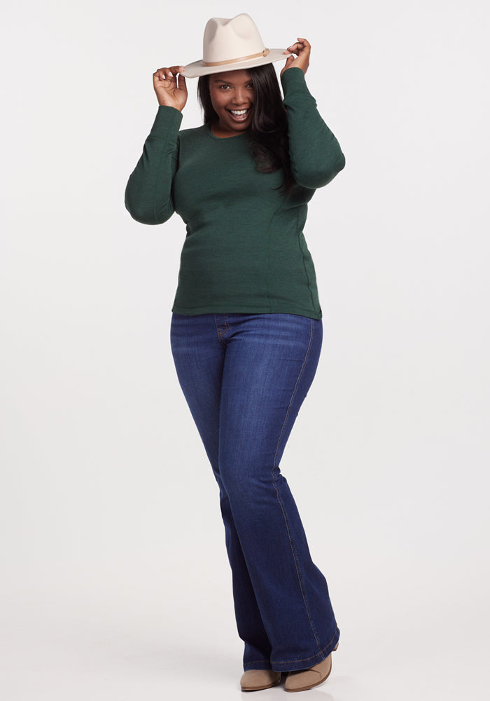 A smiling woman wears the Woolx Hannah Long Sleeve Top in Forest over an odor-resistant, moisture-wicking Merino wool base layer, paired with blue flared jeans, beige boots, and a light hat, holding the brim with both hands against a white background.