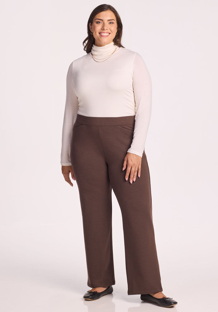 A woman smiles while standing against a plain light background, wearing a white turtleneck, the Woolx Ellie Wide Leg Pants in Bracken, black flats, and a layered gold necklace.