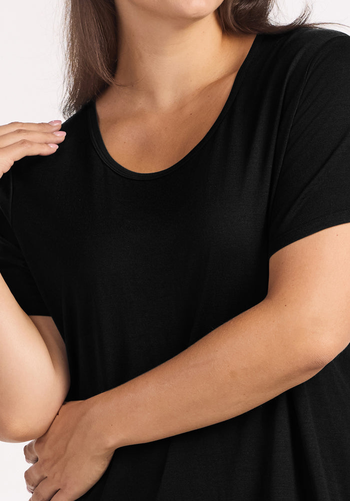 A woman wears the Woolx Desi Nightgown in black, posing with one hand near her neck and the other on her arm. The cropped image captures her from shoulders to mid-torso.