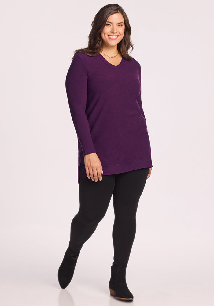 A woman with long brown hair smiles in the Woolx Evie Ribbed Tunic - Majestic Purple, black leggings, and ankle boots. She stands against a plain light background, enjoying the comfort of this Merino wool top.