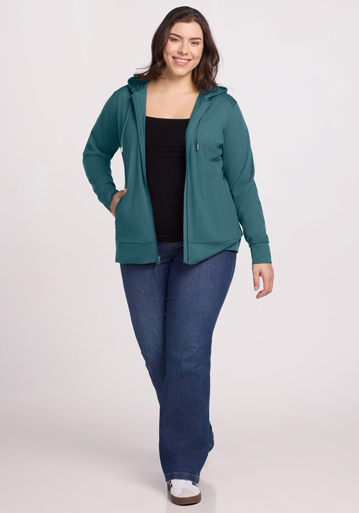 A woman with long brown hair wears the Woolx Ryann Hoodie in Tidepool over a black top and blue jeans. She stands smiling, facing forward with one hand in her pocket against a plain light background.