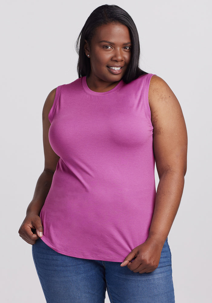 A woman with long dark hair, wearing the Woolx Bella Tunic Top in Purple Wine and blue jeans, stands against a plain light background, smiling confidently at the camera.
