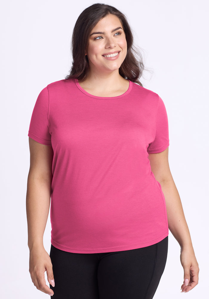 A woman with long brown hair smiles while wearing the Woolx Liza Crew Neck Tee in Raspberry Fizz and black pants, standing against a plain white background. 