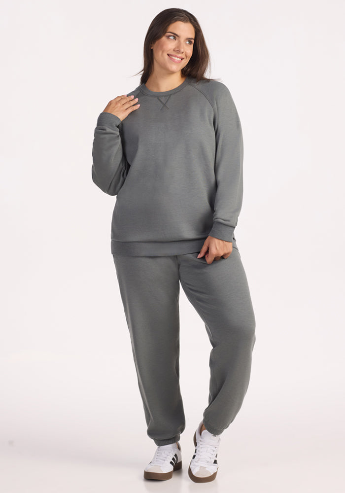 A woman smiles and touches her hair while wearing the Woolx Bailey Crewneck Sweatshirt in Sedona Sage, paired with matching sweatpants and white sneakers with black stripes. The background is plain white.