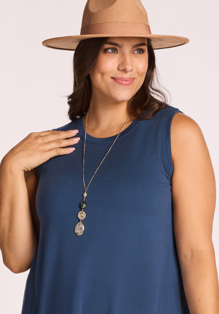 A woman smiles while looking to the side, resting one hand on her chest as she wears the Woolx Clara Dress - Starry Night, paired with a tan wide-brimmed hat and a long charm necklace.