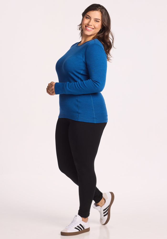 A woman with long brown hair smiles while wearing the Woolx Hannah Long Sleeve Top in Nightfall, black leggings, and white sneakers with black stripes. She stands sideways against a white background, highlighting her outfit’s temperature-regulating features.
