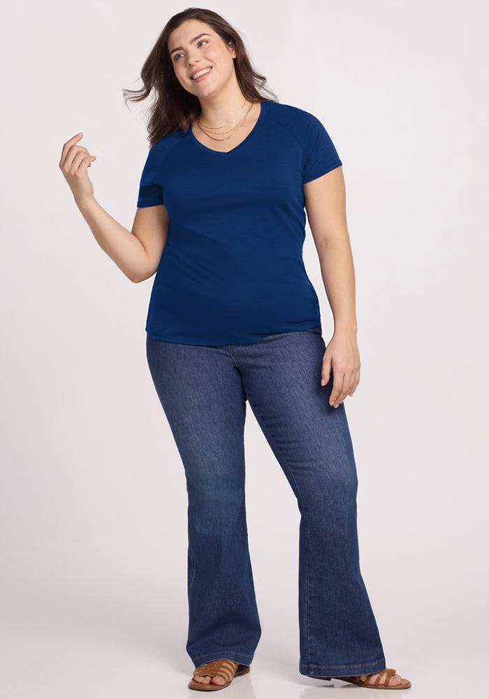 A woman wearing the Woolx Mia V Neck in Estate Blue, bootcut jeans, and brown sandals stands smiling against a plain light background.