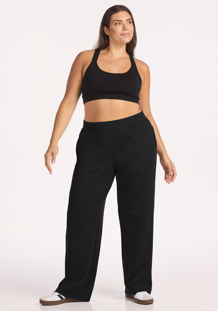 A woman with long brown hair and a relaxed, confident look models the Woolx Bree Lounge Pants in black against a plain white background, paired with a black sports bra and white sneakers.