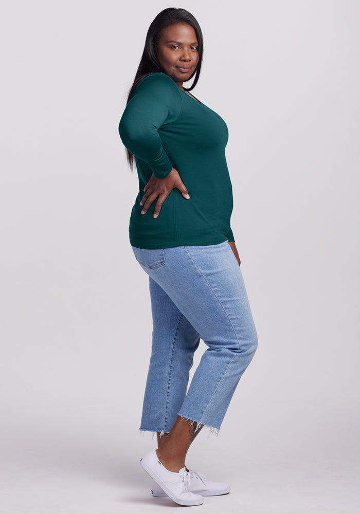 A woman stands sideways, looking at the camera with one hand on her hip. She wears the Woolx Remi Long Sleeve T-Shirt in Riverbank Green, light blue cropped jeans with frayed hems, and white sneakers against a plain light background.
