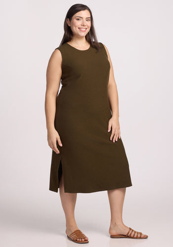 A person confidently poses in a sleeveless Iris Ribbed Dress by Woolx in an olive leaf shade, featuring side slits. They wear brown sandals and have long dark hair against a plain, light backdrop. | Juliana is 5'10", wearing a size XL