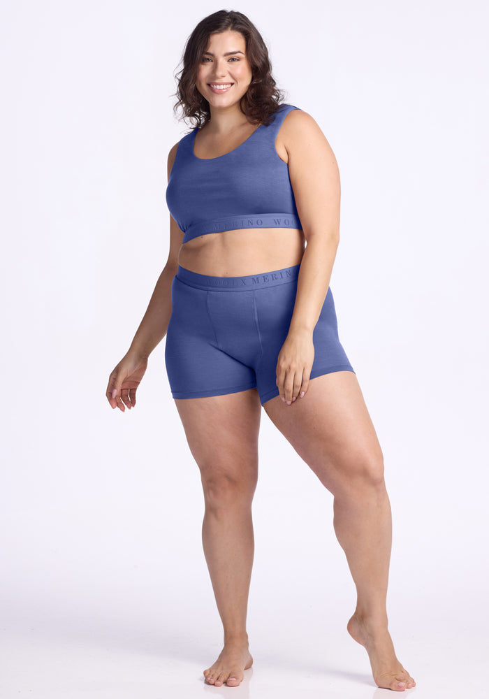 A smiling woman with wavy brown hair wears the Woolx Elliott Boyshort in Cloudy Bay and a matching sports bra, standing barefoot against a plain white background.