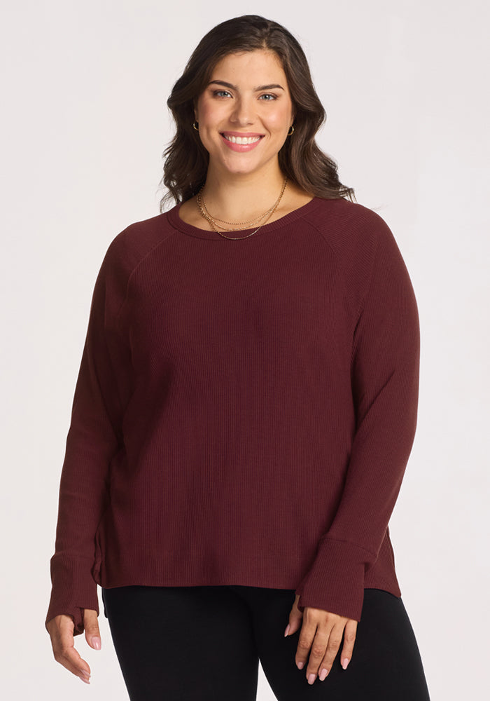 A woman with long brown hair, wearing the Woolx Ainsley Sweater in Toasted Garnet and black pants, smiles while standing against a plain light background. 