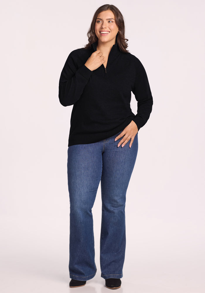 A woman smiles confidently, wearing the Woolx Collins Half-Zip Sweater in black with blue jeans and dark shoes. She poses against a plain light background, one hand near her neck and the other on her hip.