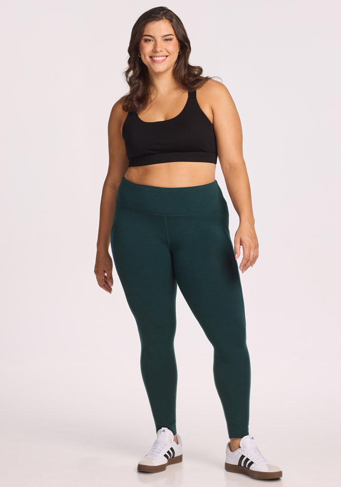A woman with long brown hair, wearing a black sports bra and Woolx Piper Pocket Leggings in Deep Timber, smiles against a plain light background. She also sports white sneakers with black stripes.