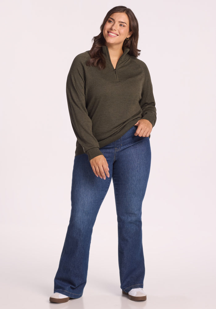 A woman smiles while posing in the Woolx Collins Half Zip Sweater in Pine Needle and blue bootcut jeans with white sneakers against a plain light background.
