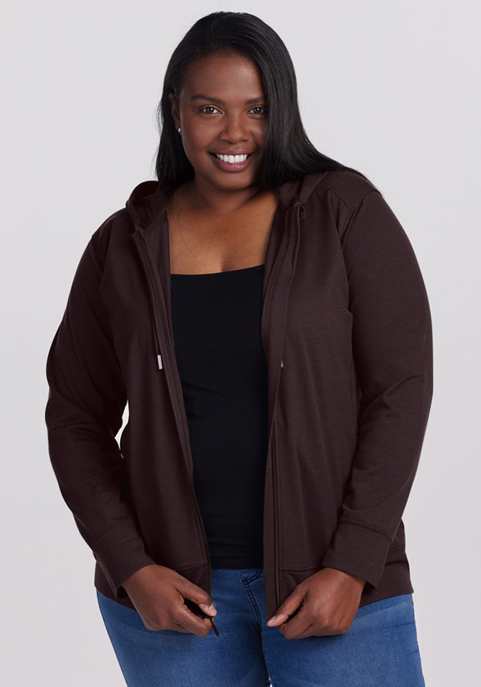 A woman with long dark hair smiles while wearing a black top, blue jeans, and the Woolx Ryann Hoodie in French Roast against a plain light background. 