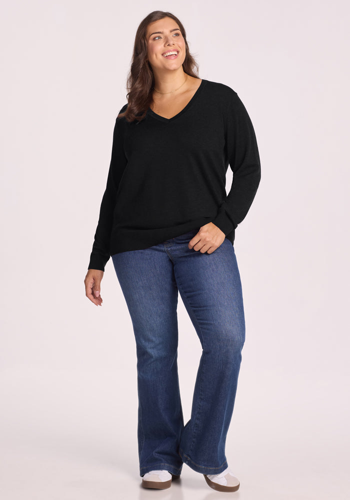 A woman with long brown hair smiles against a plain light background, wearing the Woolx Francesca Sweater in black, paired with blue flared jeans and white shoes.