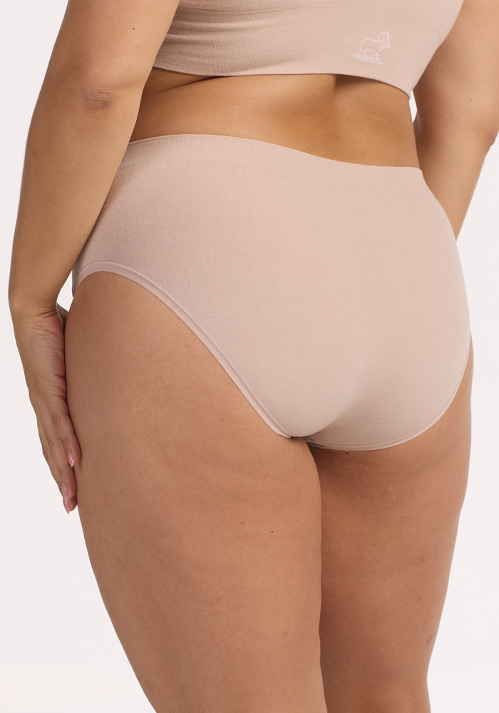 A person wearing the Woolx Maeve Bikini in Sand—seamless, high-waisted underwear made from moisture-wicking fabric—with a matching bra, shown from the back and resting one hand on their hip.
