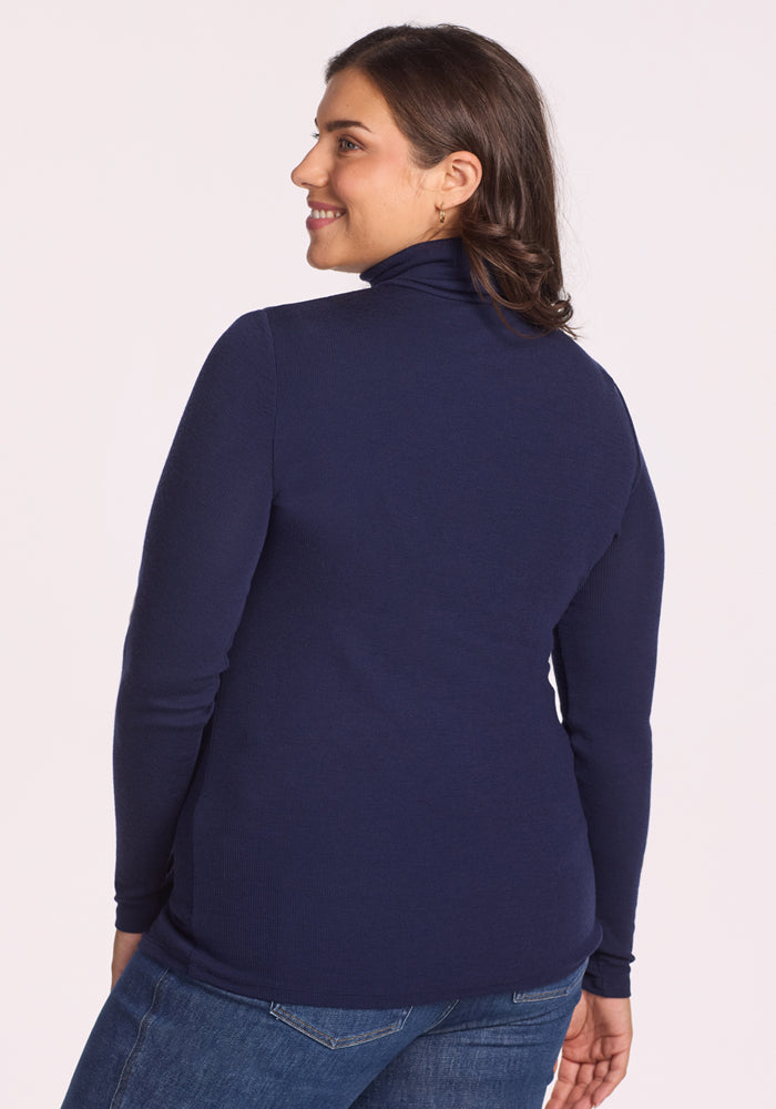A woman with shoulder-length brown hair smiles over her shoulder, wearing the Woolx Sage Ribbed Turtleneck in Midnight Sapphire and blue jeans, standing against a plain light background.