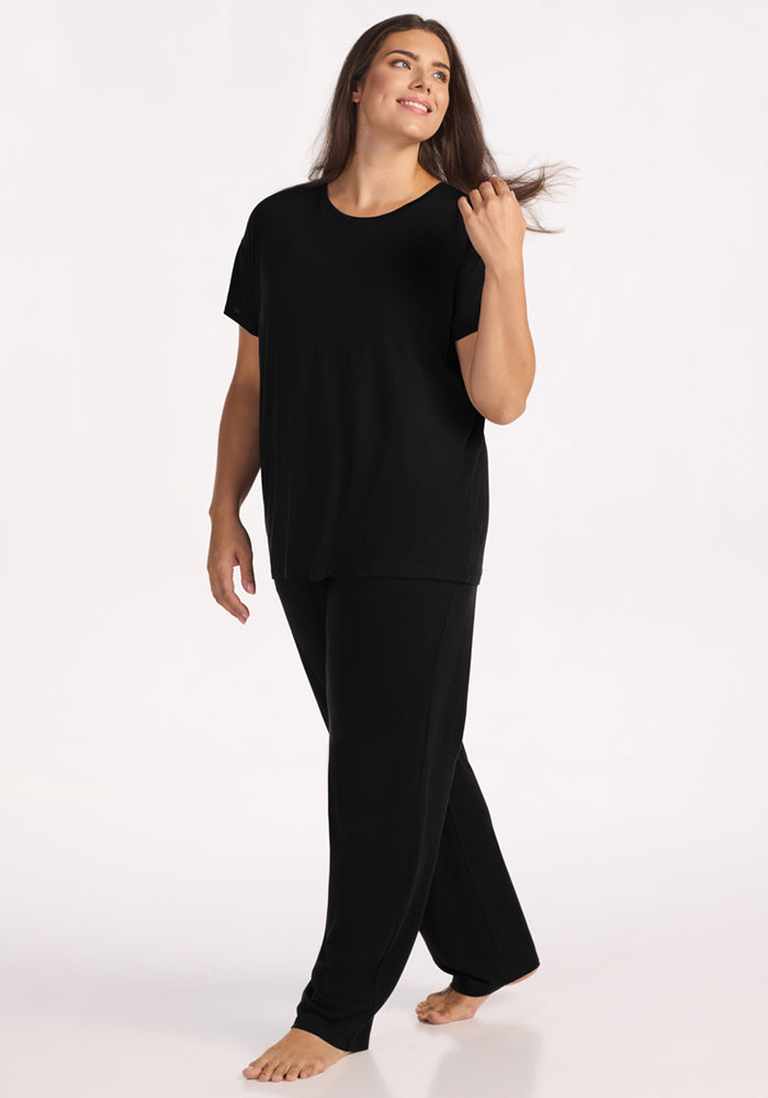 A woman with long brown hair, barefoot and smiling, walks forward while touching her hair. She wears the Woolx Billie Lounge Tee in black with matching pants, set against a plain light background.