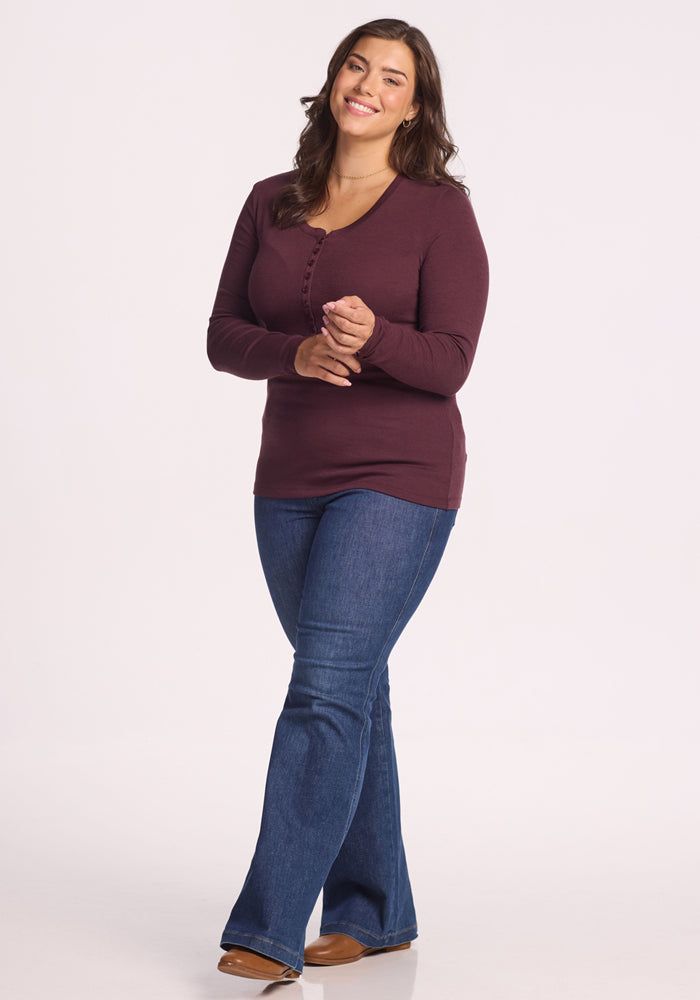 A woman with long brown hair smiles against a plain background, wearing the Woolx Reese Ribbed Henley in Chocolate Fig, blue jeans, and brown shoes, with her hands clasped in front.