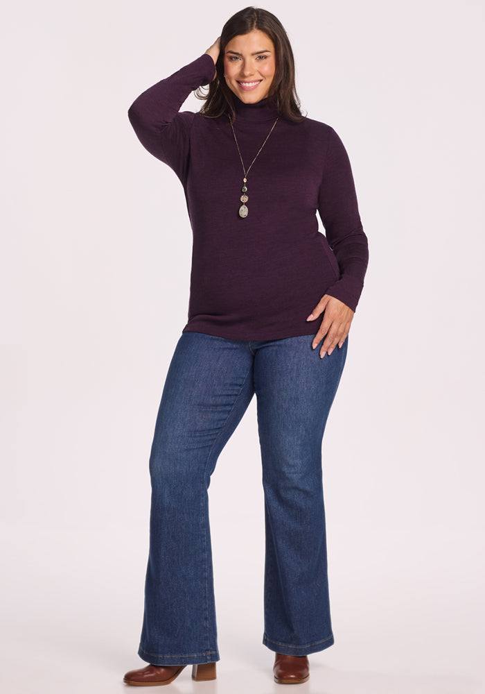 A woman with long dark hair smiles, standing with one hand on her head and the other on her hip. She wears the Woolx Peyton Turtleneck in Deep Plum, blue jeans, brown heeled boots, and a long pendant necklace.