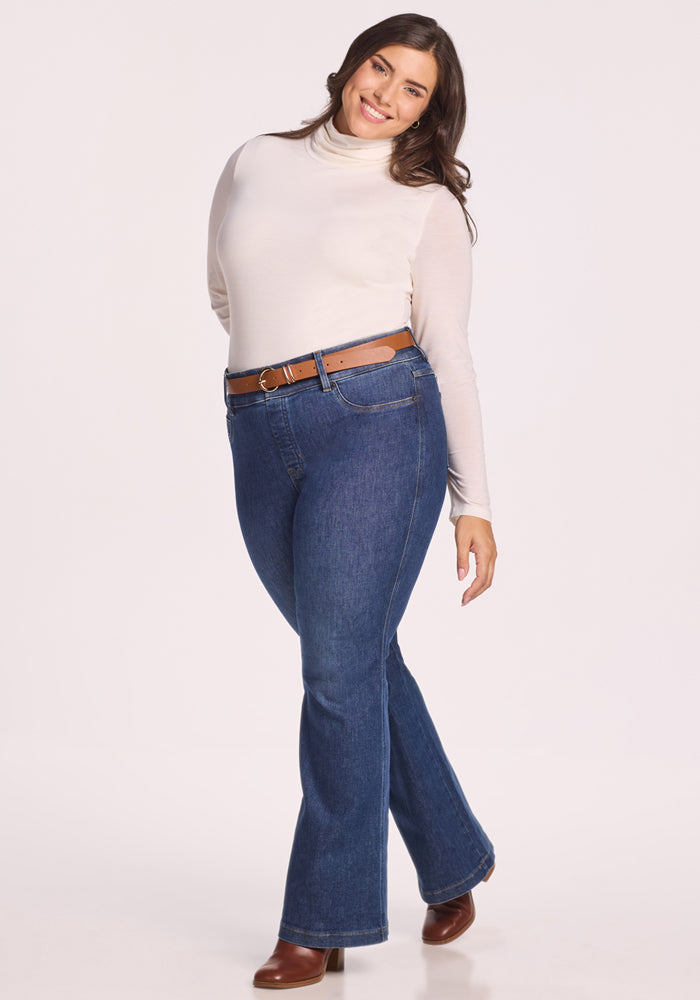 A woman smiles and poses in the Woolx Kennedy Bodysuit - Buttercream, styled with blue jeans, a brown belt, and brown boots against a plain light background. This must-have piece brings timeless style to her look.