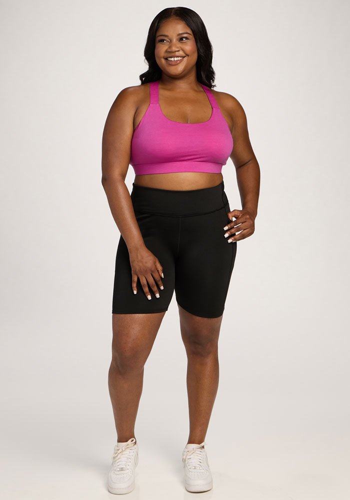Woman smiles in the Woolx Livi Bra - Summer Sorbet with black bike shorts and white sneakers against a plain background.