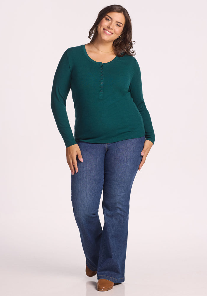 A woman with long brown hair smiles while wearing the Woolx Reese Ribbed Henley in Riverbank Green, paired with blue jeans and brown shoes, standing against a plain light background.