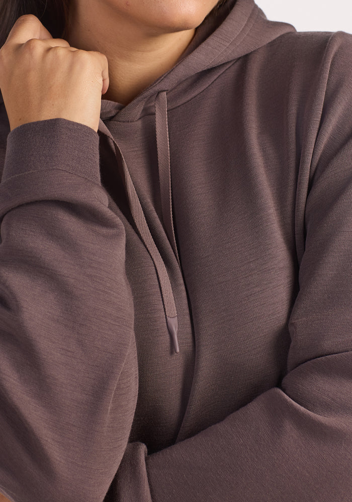 A person wearing the Woolx Avery Hoodie in Plum Truffle with visible drawstrings, resting their chin on their fist and crossing their other arm over their chest. Only the lower half of their face is shown.